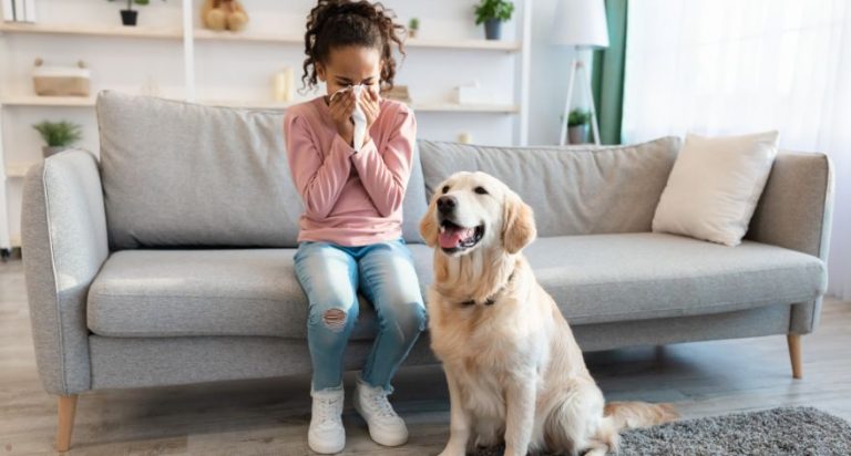 A teenager sitting on a couch sneezing into a Kleenex. A golden retriever is sitting on the floor beside her.