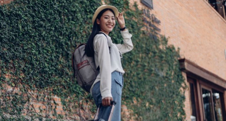 A woman standing in front of a brick wall covered in ivy. She has a suitcase and a backpack.